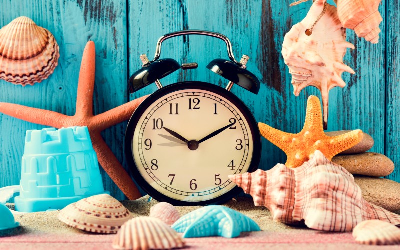 an alarm clock being adjusted forward one hour at the beginning of the summer time, many conches and starfishes, and a beach pail, on a pile of sand, against a bright blue rustic wooden background