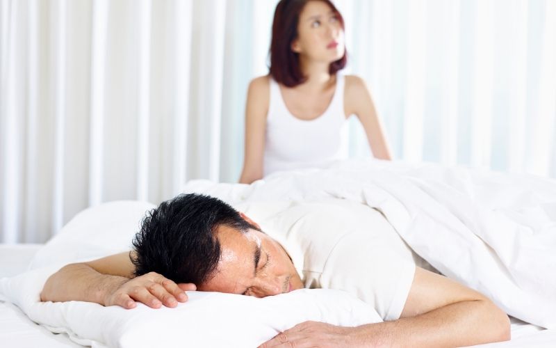 husband sleeping in bed while wife looks worried