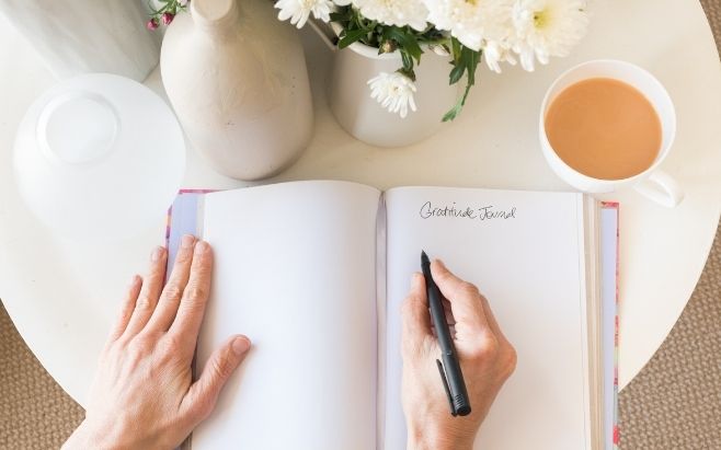 woman writing in gratitude journal on table with coffee and flowers