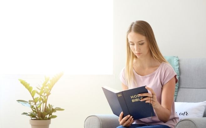 woman reading Holy Bible and sitting in couch with potted plant next to her