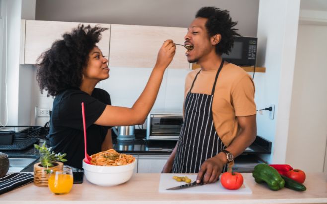 woman serving man a spoon with food in kitchen while cooking