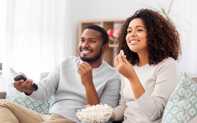 man and woman sitting on couch eating popcorn watching tv