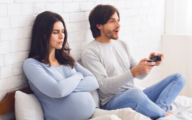 upset pregnant woman sitting on bed next to man playing video games