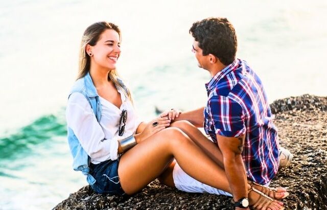 couple sitting on beach rock smiling and holding hands